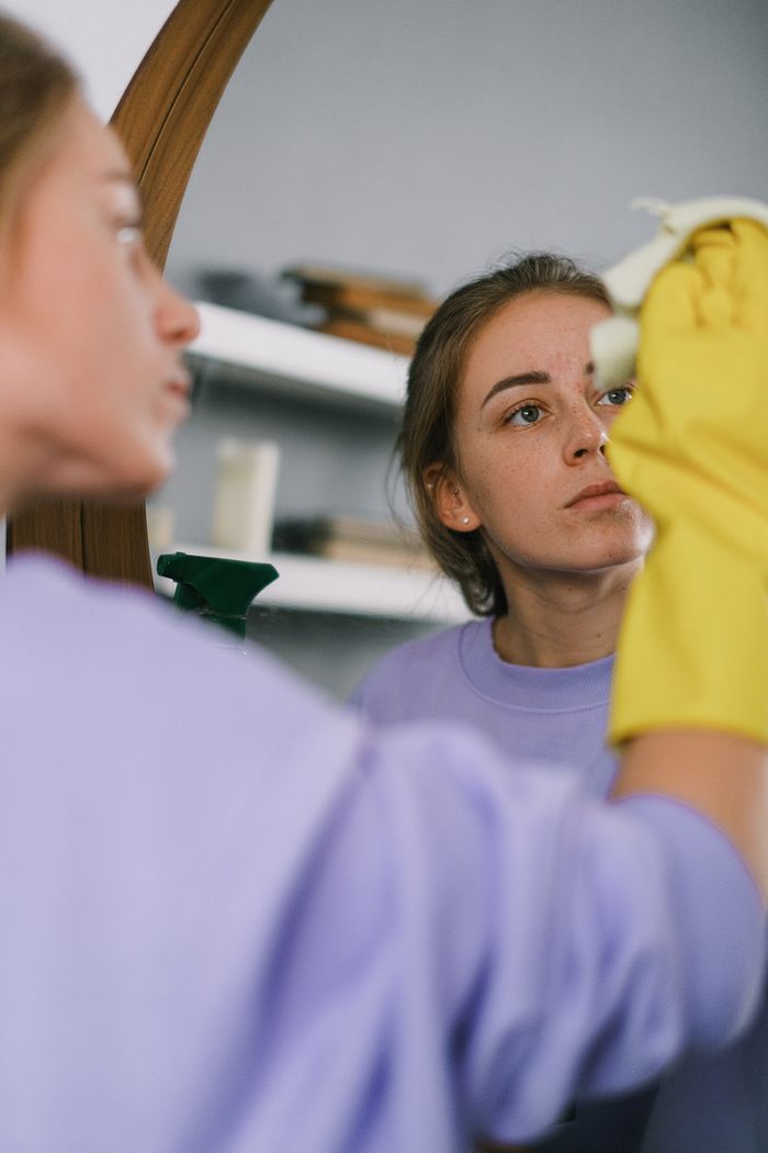about-us-02 Crop female in purple sweatshirt and latex gloves washing mirror in light apartment in daytime